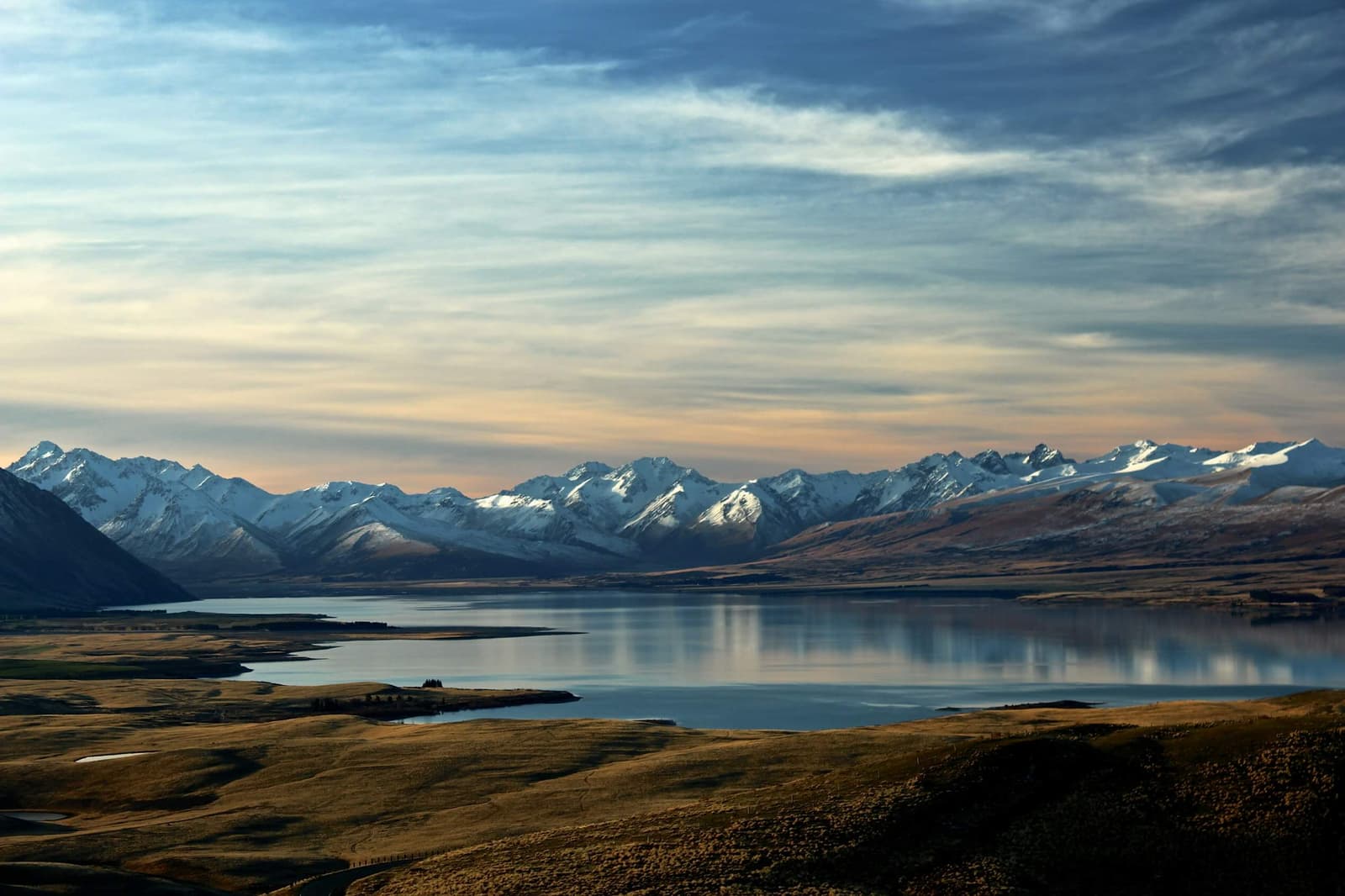 Vista de umas pequenas colinas, um grande lago e montanhas com neve ao fundo