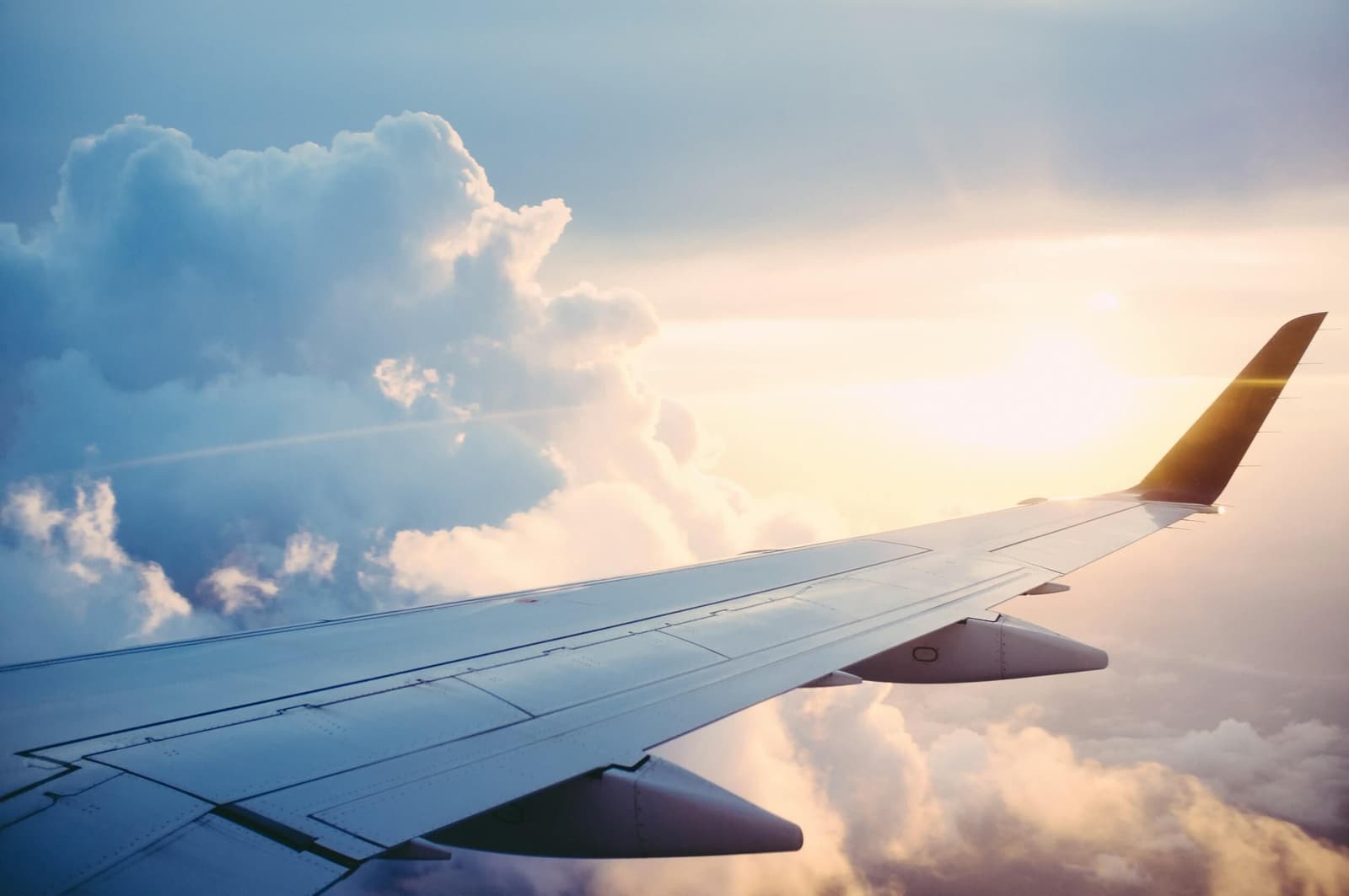 Vista da asa de um avião durante o voo acima das nuvens ao pôr do sol, com o céu iluminado em tons dourados e azuis suaves